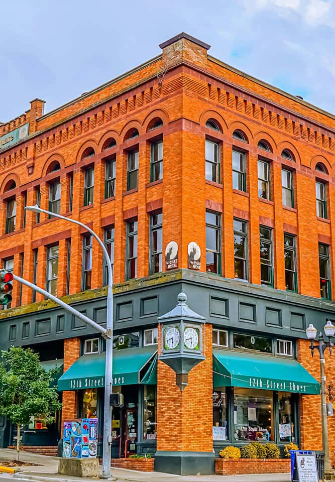 A historic red-brick building with large arched windows and green awnings houses a shoe store called 12th Street Shoes. A street clock and traffic lights are visible at the corner.