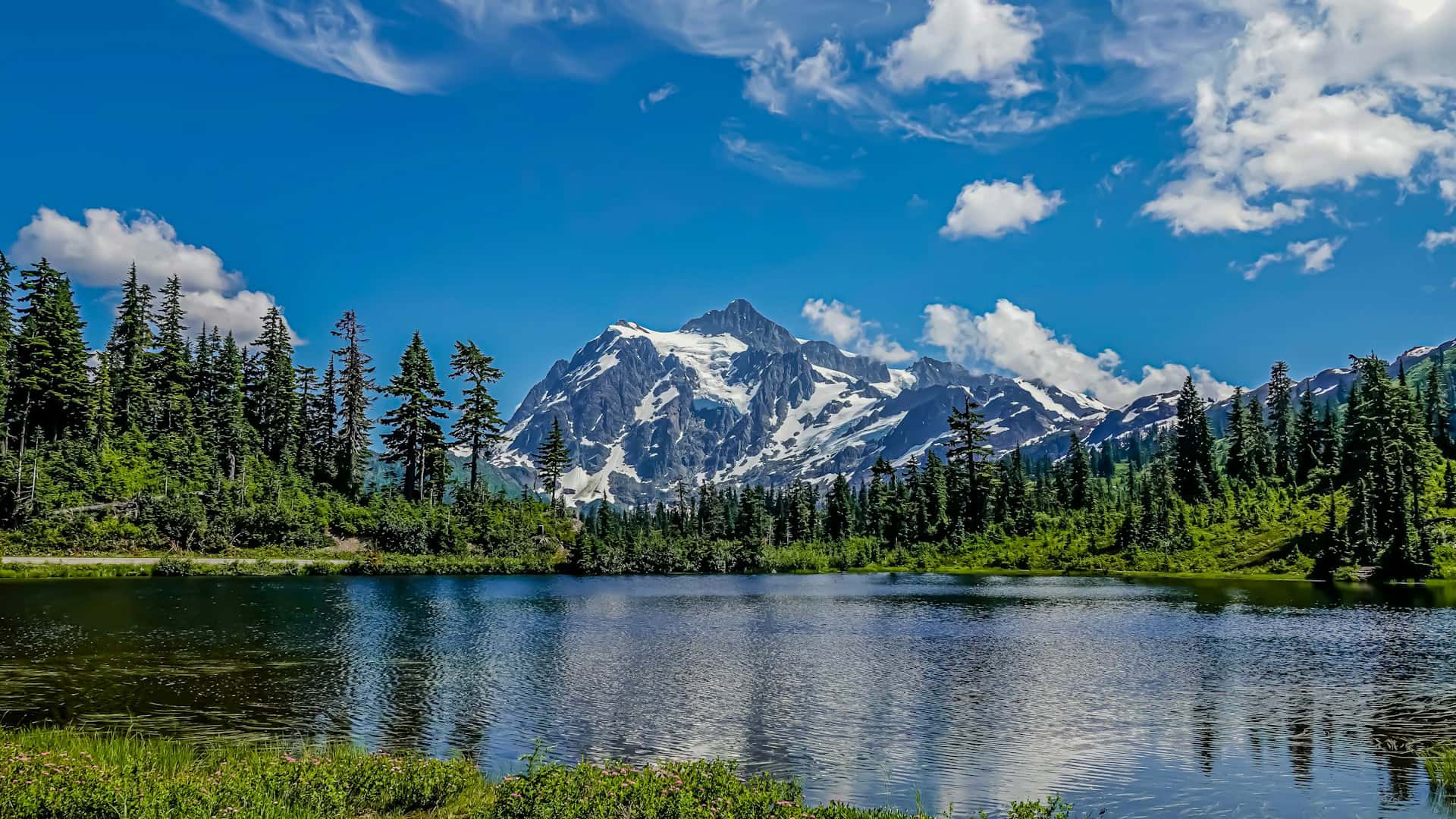 A clear lake reflects green trees and a snow-capped mountain beneath a blue sky with scattered clouds.