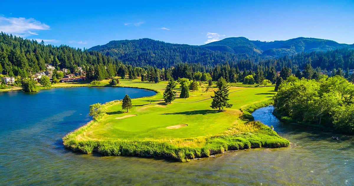 A scenic aerial view of a lush green golf course on a small peninsula surrounded by a river and lake, bordered by dense forest and distant hills under a bright blue sky.